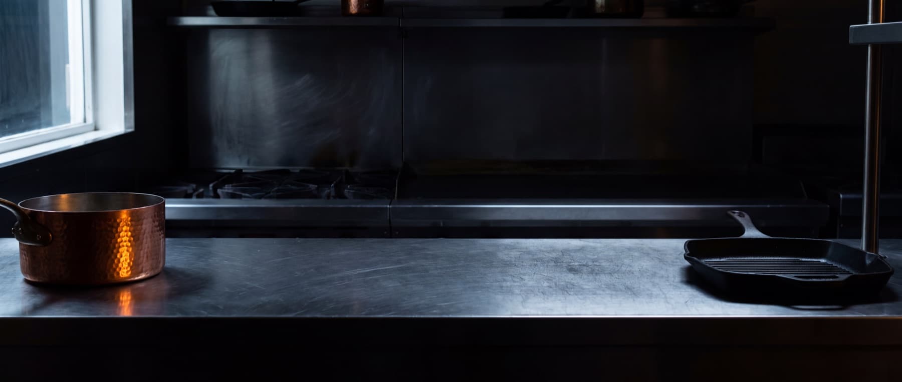Long plan de travail en inox brossé la nuit, un faitout en cuivre martelé et un gril en fonte posés loin l'un de l'autre, une lame de lumière froide venant de la gauche