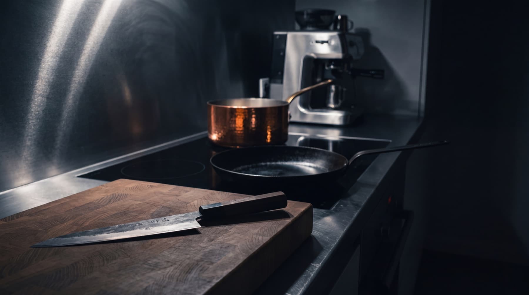 Plan de travail en inox la nuit : un couteau de chef posé sur un billot en bois de bout, une poêle en fer noire, un faitout en cuivre et une machine expresso en arrière-plan